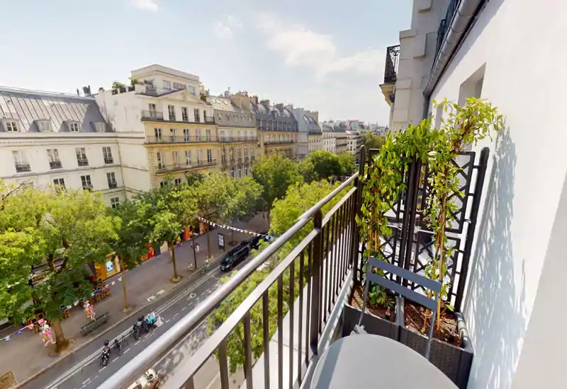 Balcón soleado con barandilla metálica, macetas y una mesa pequeña — ofrece una agradable vista a una calle arbolada de estilo parisino.
