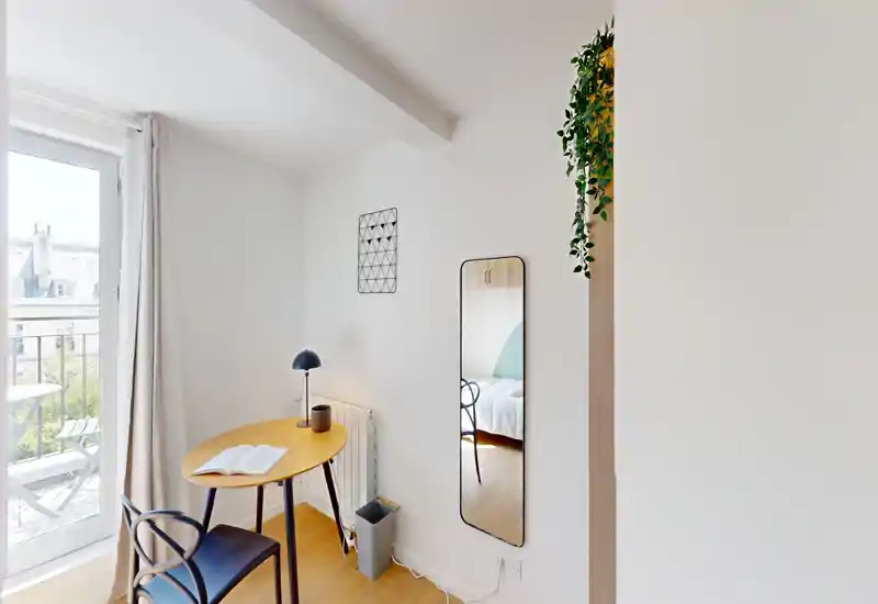 Rincón de dormitorio luminoso y minimalista con una pequeña mesa de trabajo, silla y un espejo que refleja parte de la cama; perfecto para una persona o estancias cortas.