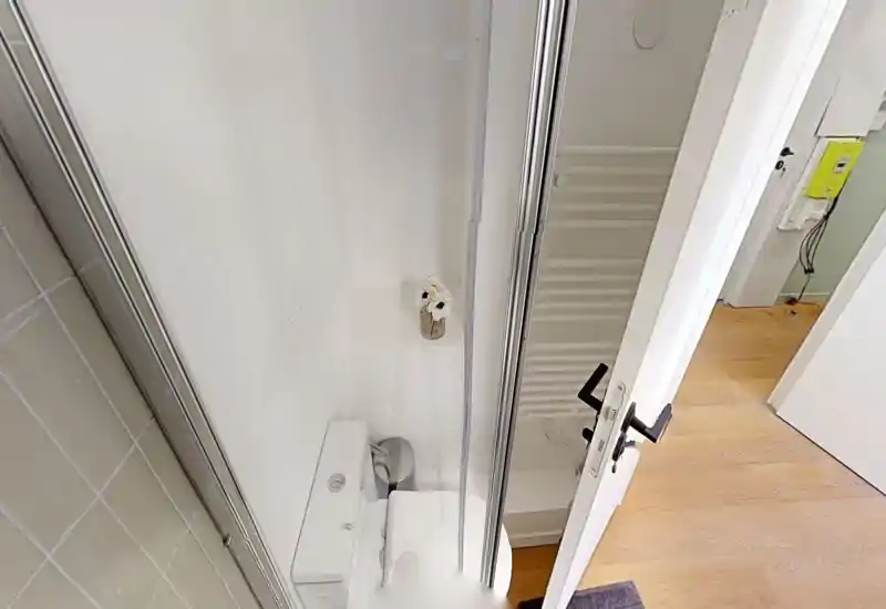 Baño compacto con inodoro y cabina de ducha, azulejos blancos limpios y puerta de ducha de cristal; se ve el suelo de madera del pasillo fuera de la puerta abierta.