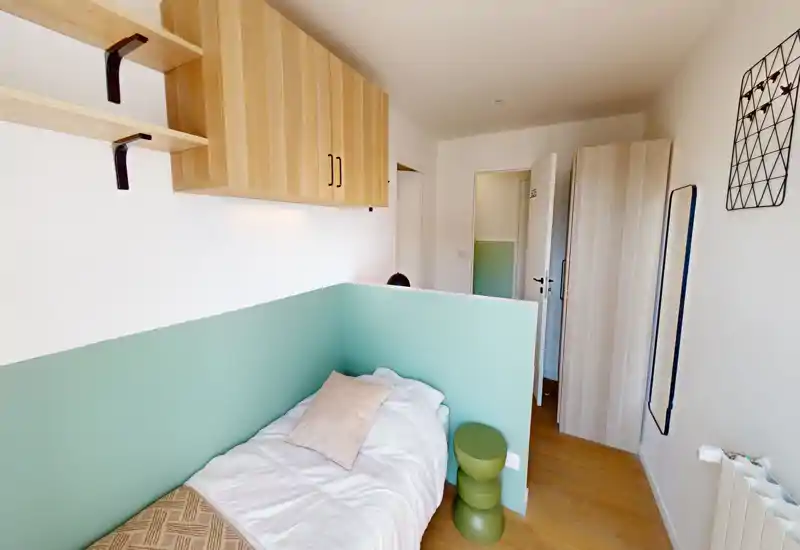 Dormitorio individual compacto y luminoso con un separador bajo verde menta, muebles altos de madera y una cama cómoda con ropa de cama suave y cojín.