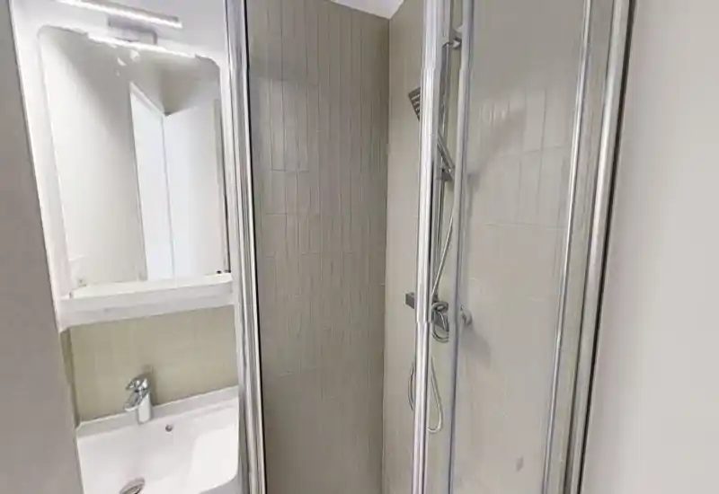 Baño compacto y moderno con ducha acristalada, azulejos en la pared, ducha de mano y lavabo pequeño con espejo: limpio y funcional, ideal para estancias cortas.