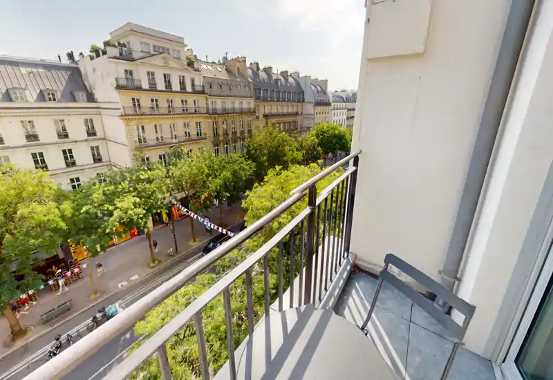 Balcon ensoleillé avec petite table et chaise donnant sur une rue bordée d'arbres et de belles façades haussmanniennes — parfait pour un café matinal avec vue sur la ville.