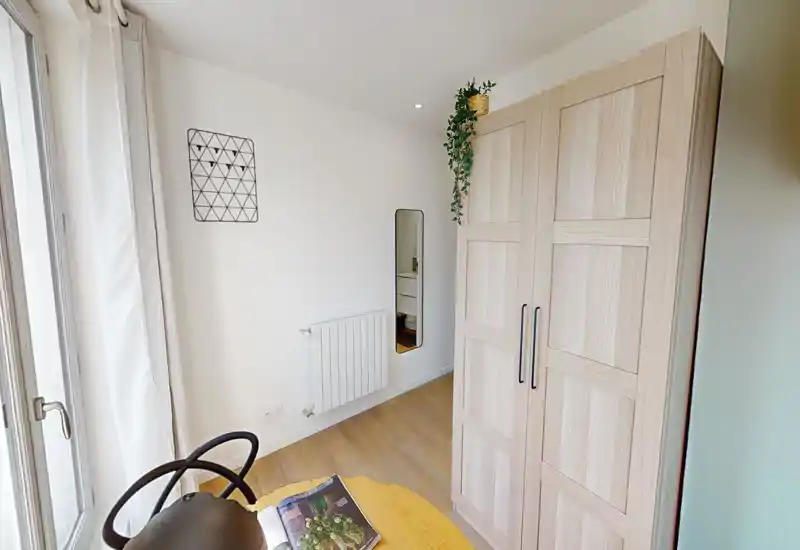 Coin de chambre compact avec une grande armoire en bois, fenêtre du sol au plafond avec rideaux légers et petite table ronde, lumière naturelle abondante et décoration scandinave épurée.