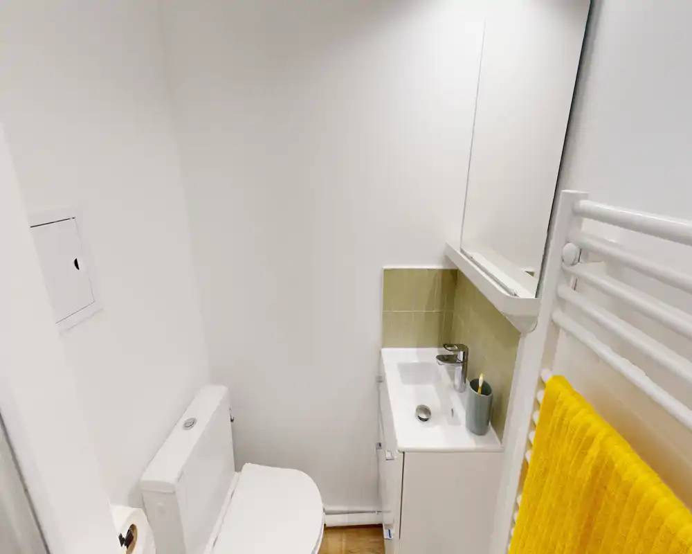 Compact, bright bathroom with a toilet, small sink cabinet and a mirror. Clean white walls and a heated towel rail with a yellow towel add a fresh, modern touch.
