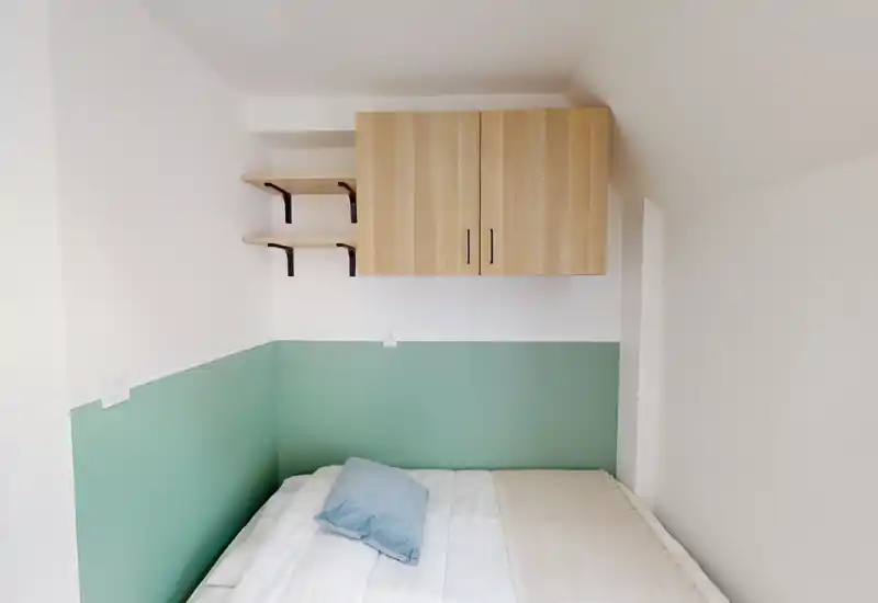 Coin chambre compact et lumineux avec un lit double adossé à une demi-cloison vert menthe, un oreiller et des rangements muraux en bois: ambiance minimaliste et chaleureuse.