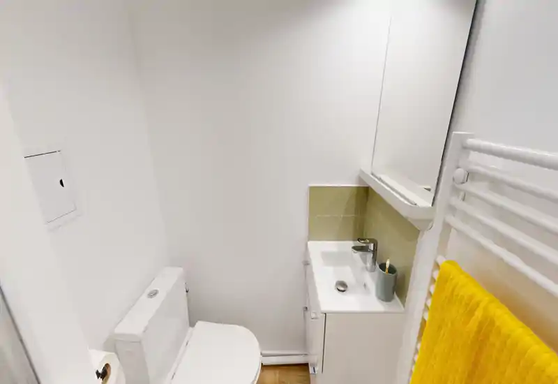 Salle de bain compacte et lumineuse avec WC, petit meuble lavabo et miroir. Murs blancs et sèche-serviettes avec une serviette jaune apportant une touche moderne et fraîche.