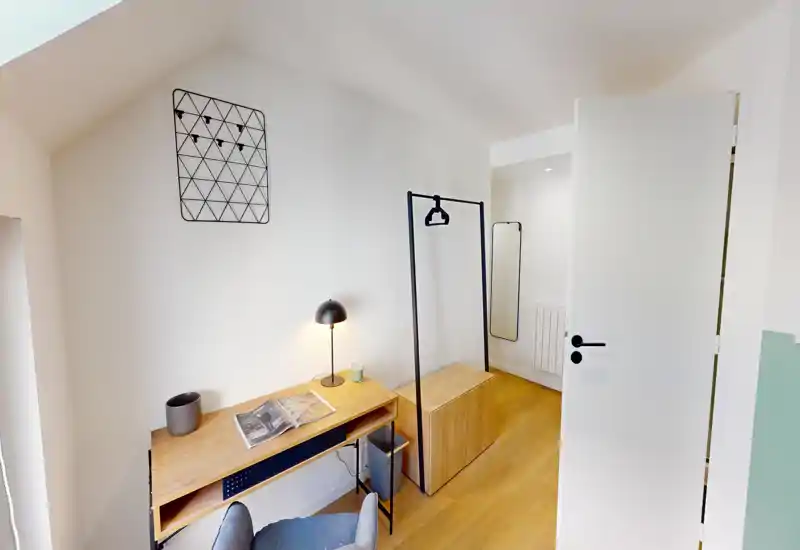 Petit espace de vie avec bureau en bois, fauteuil et porte-manteau; lumineux, minimal et adapté à une personne ou à un coin travail.