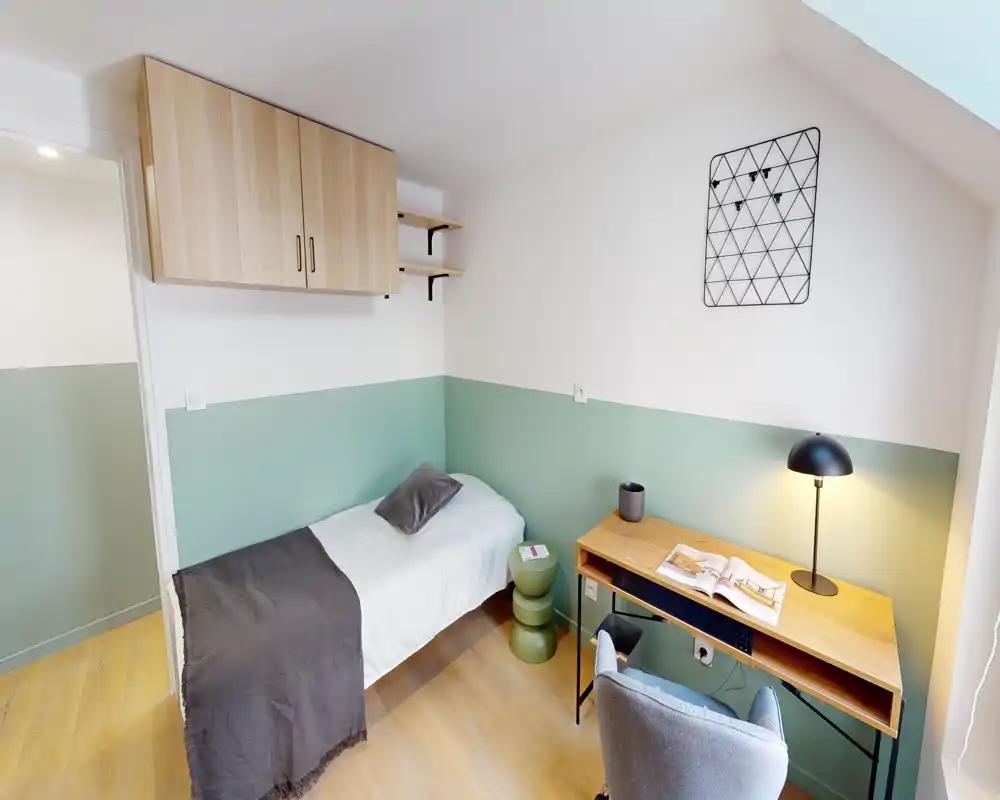 Piccola camera singola ben arredata con letto, pensili a parete e una scrivania in legno illuminata da una lampada da tavolo — ideale per uno studente o single.