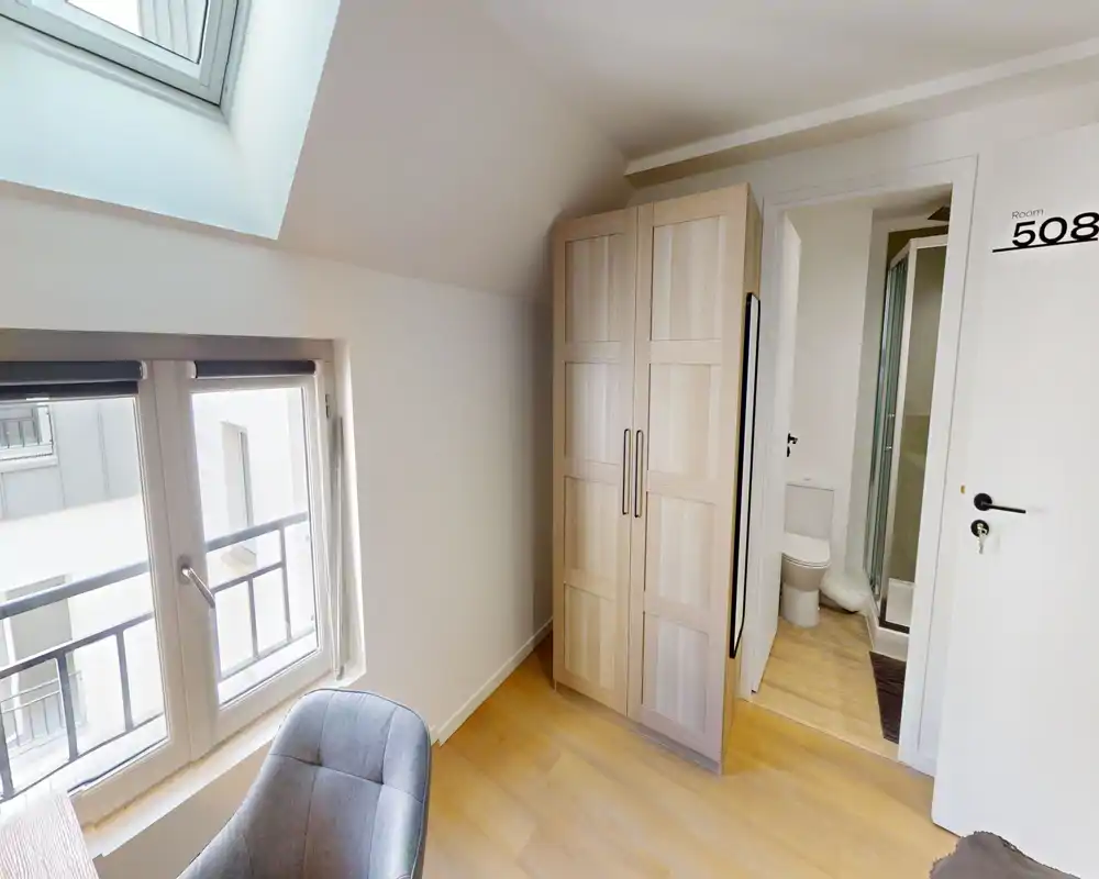 Angolo di una camera mansardata con armadio, poltroncina e bagno en-suite visibile. La luce naturale dal lucernario e dalla finestra rende l’ambiente luminoso e accogliente nonostante la metratura compatta.