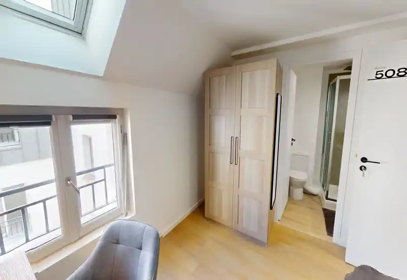 Coin de chambre mansardée avec armoire, fauteuil et salle de bains attenante visible. La lumière naturelle provenant du velux et de la fenêtre rend l’espace lumineux et accueillant malgré la compacité.
