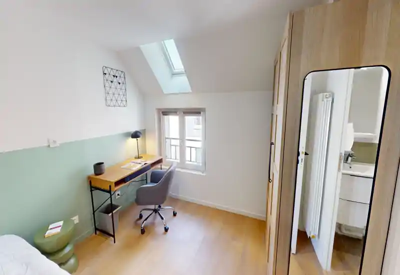 Chambre compacte et lumineuse avec coin bureau : petit lit double (partiellement visible), bureau en bois avec chaise sous une fenêtre de toit et armoire avec miroir intégral. Décor moderne et épuré, lumineuse, adaptée pour une personne ou le télétravail.