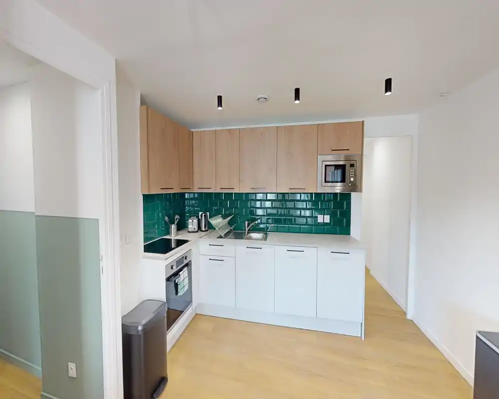 Cucina moderna compatta con basi bianche, pensili in legno e paraschizzi in piastrelle verde lucide; fornita di forno incassato, microonde, lavello e piano di lavoro ordinato — luminosa e ben organizzata per un piccolo appartamento.
