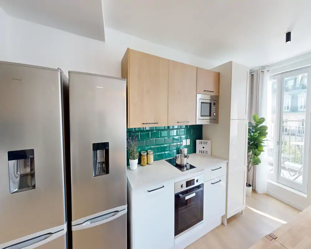 Cucina moderna e compatta con frigorifero in acciaio inox, forno incassato e rivestimento a mattoncini verdi; ambiente luminoso con luce naturale e piante decorative.