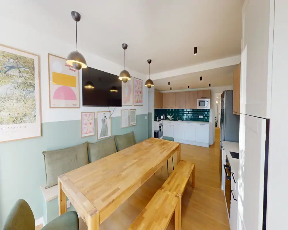 Zona pranzo moderna e luminosa con grande tavolo in legno e panche, cuscini coordinati e quadri alle pareti; layout open space con cucina contemporanea sullo sfondo.