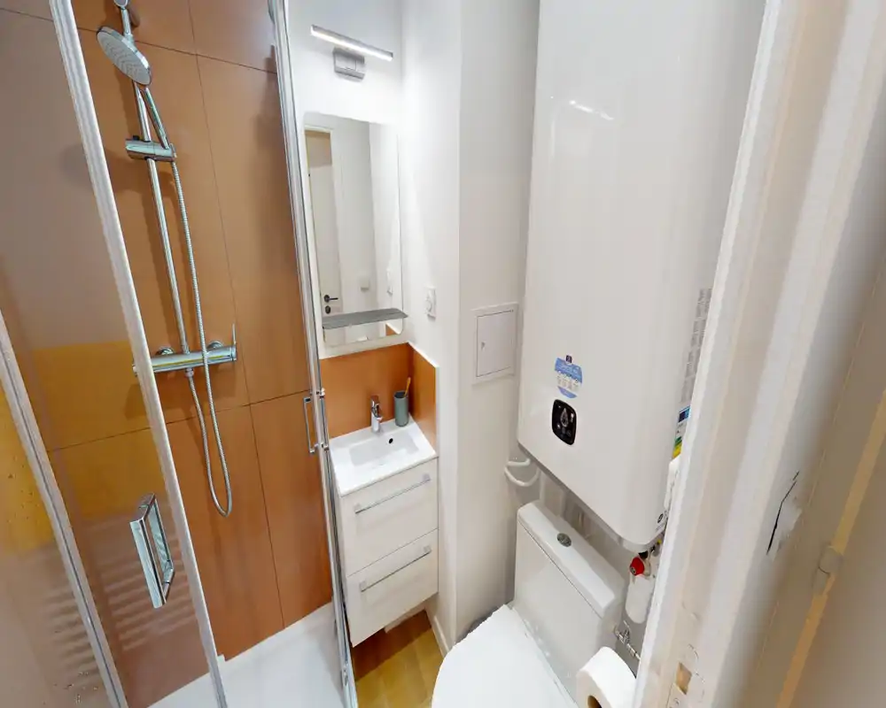 Compact, modern bathroom with a glass shower enclosure, small vanity with mirror, toilet and a mounted water heater. Clean lines and warm wood-toned shower paneling create a tidy, functional space.