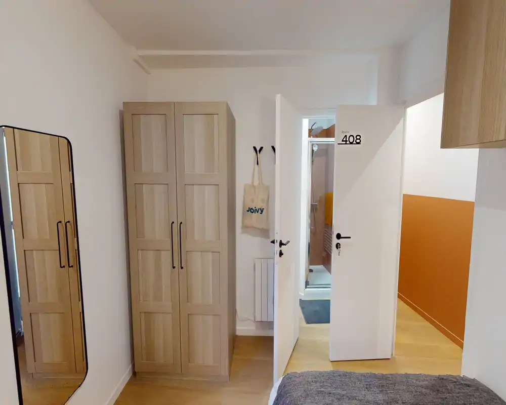 Compact bedroom with a wooden wardrobe, full-length mirror and a partially visible bed in the foreground; clean, modern styling with warm wooden flooring and neutral walls.