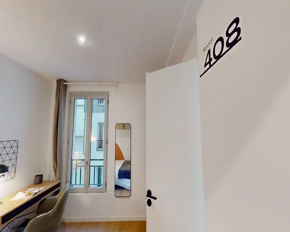 Bright, modern small bedroom seen from the doorway; a narrow mirror shows the bed, with a desk and chair by the window. Neutral décor, wooden floor and natural light create a calm atmosphere ideal for short-stay rentals.