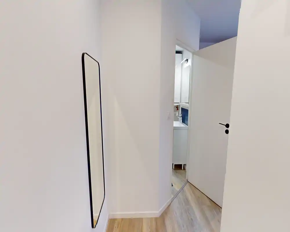 Bright, minimal entrance corridor with a full-length wall mirror and light wood flooring; clean white walls and an open door leading to a bathroom.