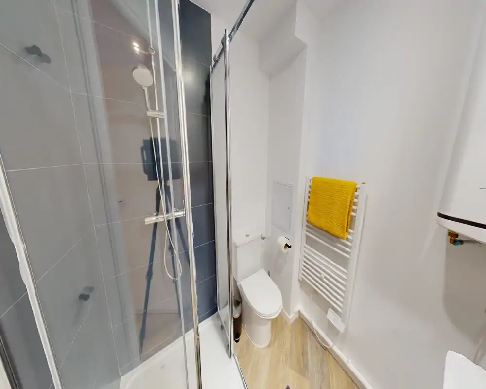 Compact modern bathroom with a glass shower enclosure, toilet and heated towel rack with a yellow towel; clean white walls and wood-effect flooring.