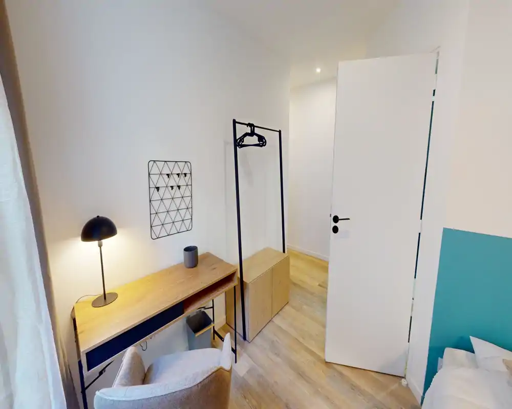 Compact, modern bedroom corner with a small visible portion of the bed, a wooden desk, lamp and open clothes rack — bright, minimal and functional.