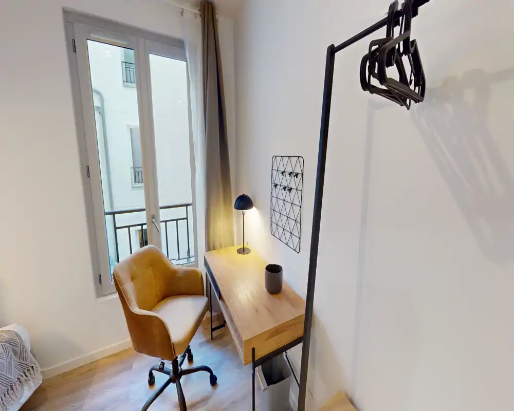 Compact bedroom corner with a small desk, warm upholstered chair and partial view of the bed by the window; bright natural light and a bedside lamp create a cozy workspace.