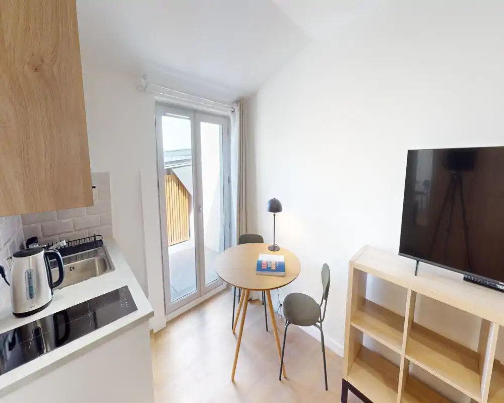 Compact kitchenette with a small dining table, two chairs and a TV. Bright natural light from the glass door and clean, modern finishes make it suitable for short-term rentals.