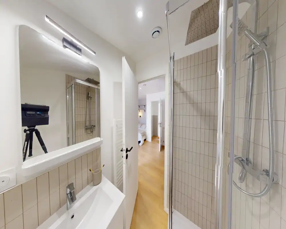 Compact modern bathroom with a wall-mounted sink, illuminated mirror and a glass-enclosed shower with chrome fixtures. The light wood floor visible through the open door leads to the bedroom, giving a neat, functional feel.