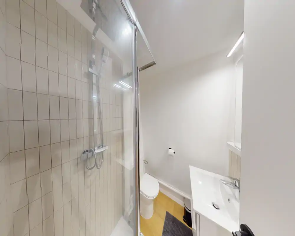 Compact modern bathroom with a glass shower enclosure, wall-mounted shower fittings, toilet, and a small sink with mirror. Bright white walls and warm wood-effect flooring give a clean, minimal look.