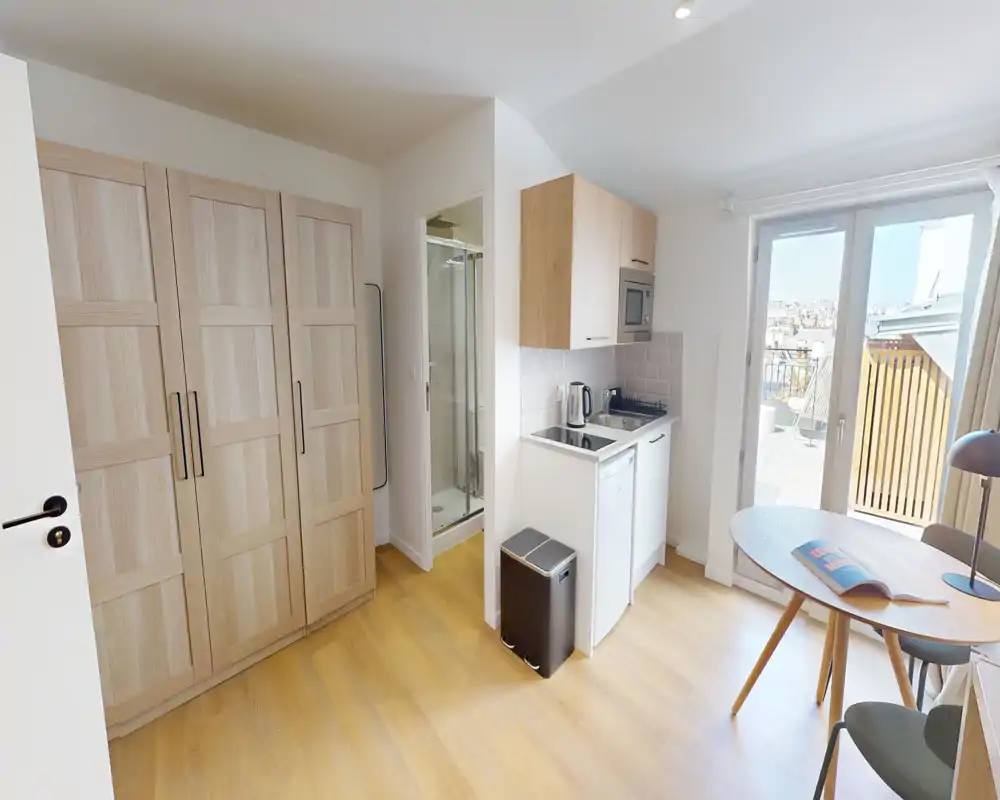 Compact kitchenette in a bright studio with light wood floors, a small dining table and direct access to a balcony. Functional appliances and storage make this a practical space for short stays.