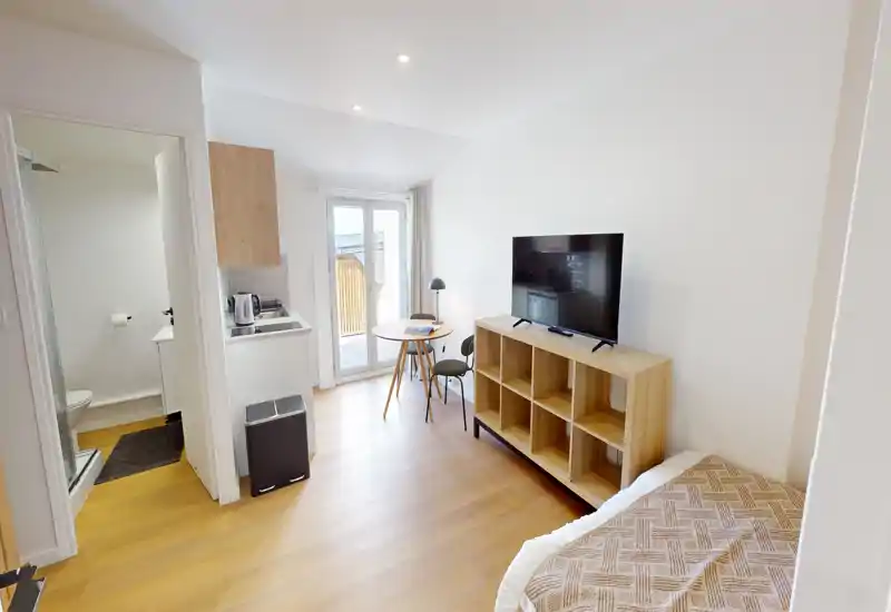 Soggiorno-studio luminoso e compatto con TV su mobile basso, piccolo tavolo da pranzo vicino alla porta del balcone, angolo cottura e bagno visibili — ideale per una persona o una coppia.