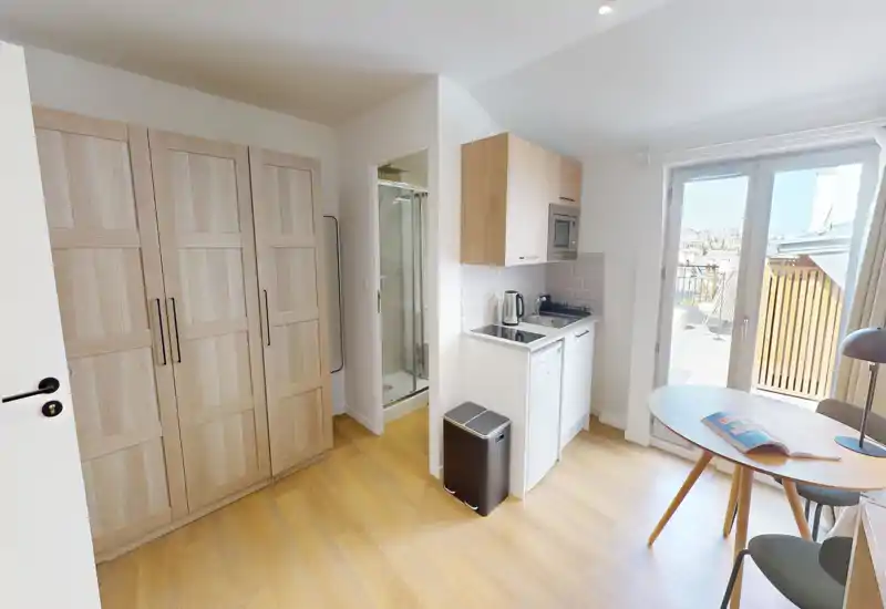 Angolo cottura compatto in un monolocale luminoso con pavimento in legno chiaro, un piccolo tavolo da pranzo e accesso diretto al balcone. Elettrodomestici funzionali e spazio di stoccaggio rendono l’ambiente pratico per soggiorni brevi.