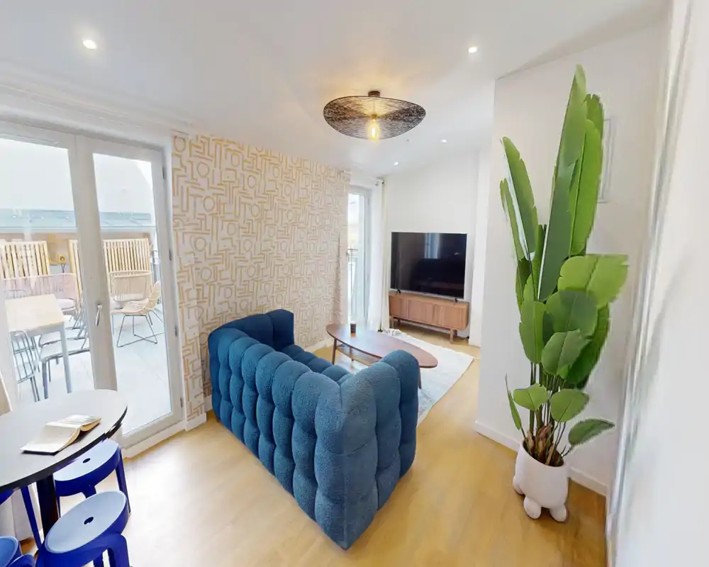 Bright, modern living room with a deep-blue tufted sofa, wooden coffee table, TV console and direct access to a balcony; tasteful wallpaper and a large potted plant complete the stylish setup.
