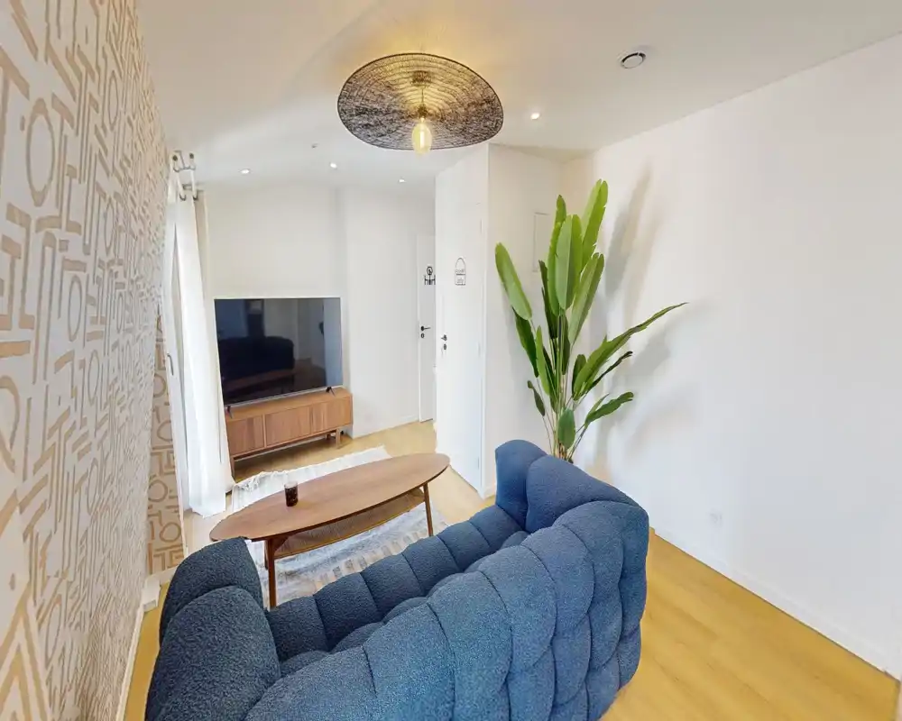 Bright, stylish living room with a tufted blue sofa, wooden coffee table and media console. Natural light and a decorative wall pattern create a warm, inviting space.
