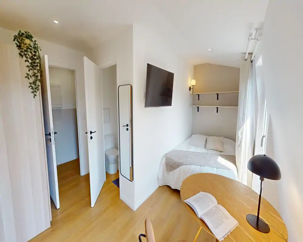 Camera da letto luminosa e ben allestita con letto matrimoniale contro la parete, TV a muro, scrivania vicino alla finestra e un piccolo bagno en-suite visibile dalla porta aperta.