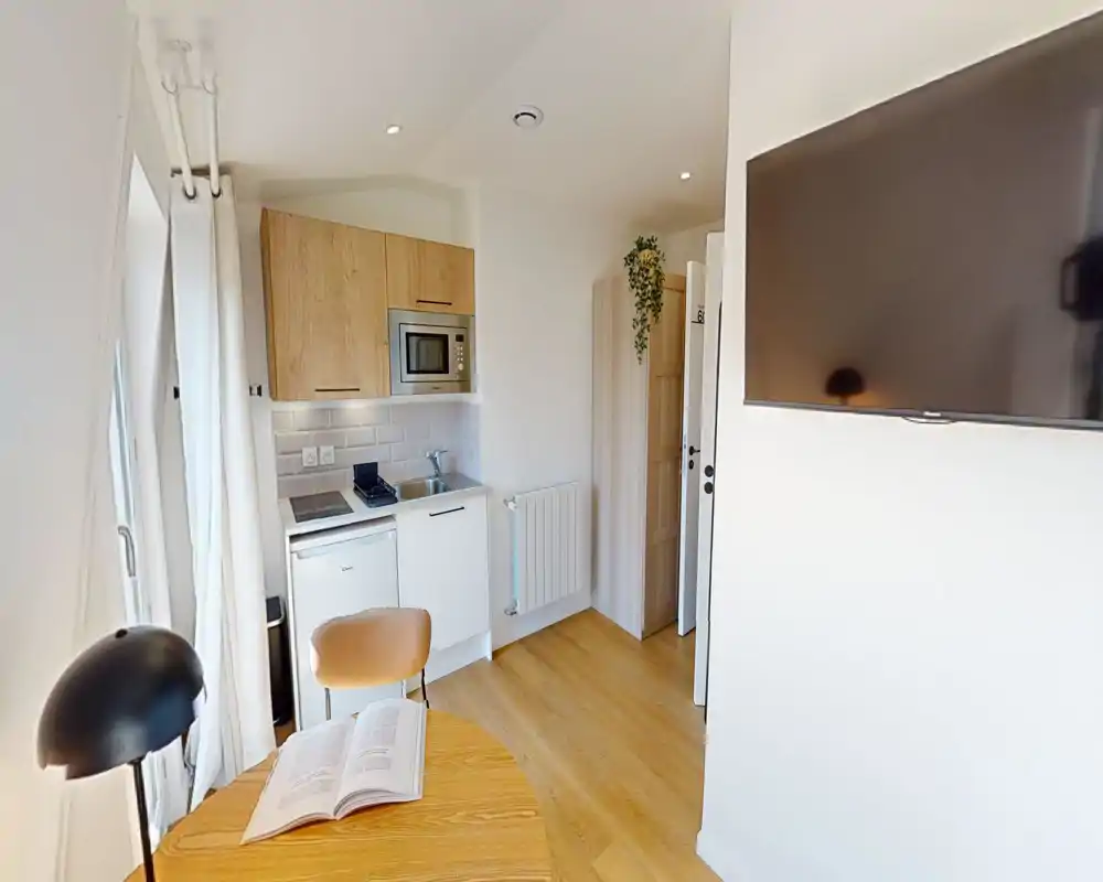 Cucinotto compatto con piccolo tavolo, TV a parete e elettrodomestici integrati (microonde, lavabo, mini-frigo). Finishes moderni e luminosi con tonalità calde del legno e finestra che garantisce luce naturale.