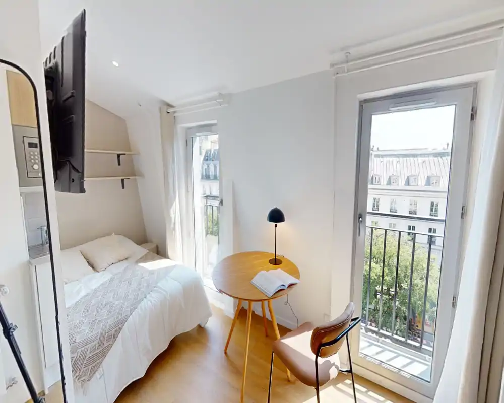 Camera da letto luminosa e compatta con letto matrimoniale, tessili accoglienti e un piccolo tavolo rotondo vicino alle grandi finestre con vista sulla città.