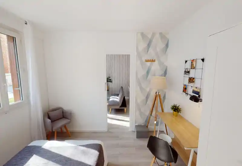 Camera da letto luminosa e moderna con il letto in primo piano, una poltroncina accanto alla finestra e una scrivania in legno con lampada decorativa — arredamento pulito in stile nordico e molta luce naturale.
