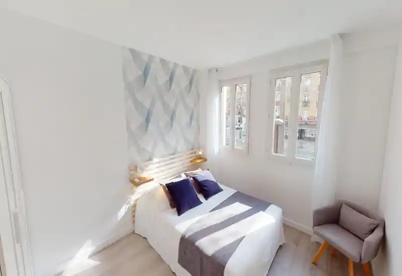Camera da letto luminosa e ben allestita con letto matrimoniale, cuscini decorativi e una parete testiera con motivo. Le grandi finestre garantiscono molta luce naturale e una poltroncina crea un angolo accogliente.