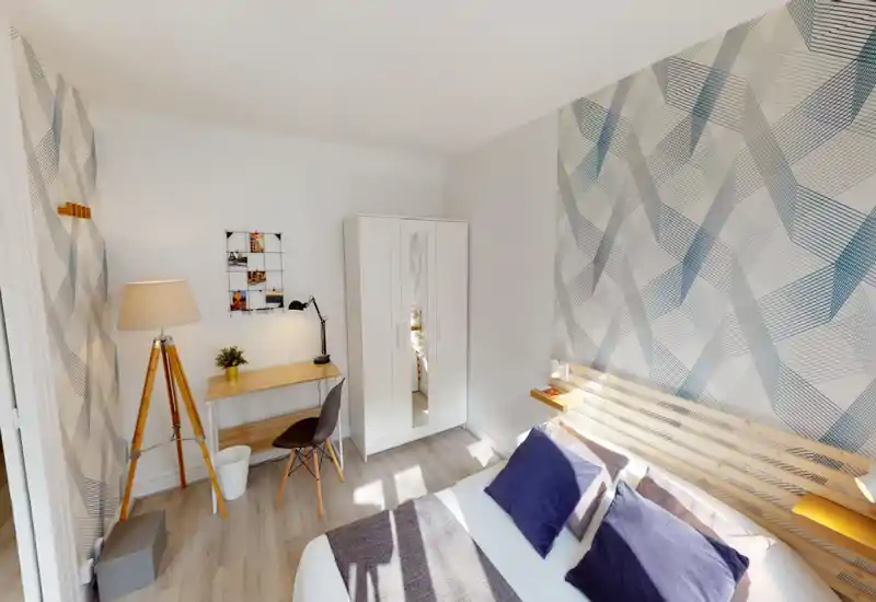 Camera da letto luminosa e moderna con ampio letto, parete decorativa e una piccola area studio. Testiera in legno, cuscini blu scuro e molta luce naturale rendono l’ambiente accogliente.