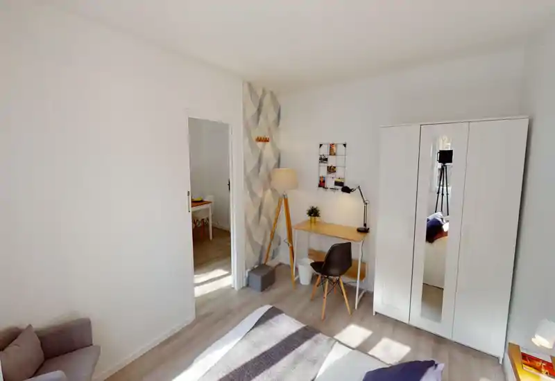 Camera da letto moderna e luminosa con letto comodo, armadio con anta a specchio e una piccola scrivania con lampada a treppiede — perfetta per una persona o coppia che cerca una stanza ordinata e soleggiata.