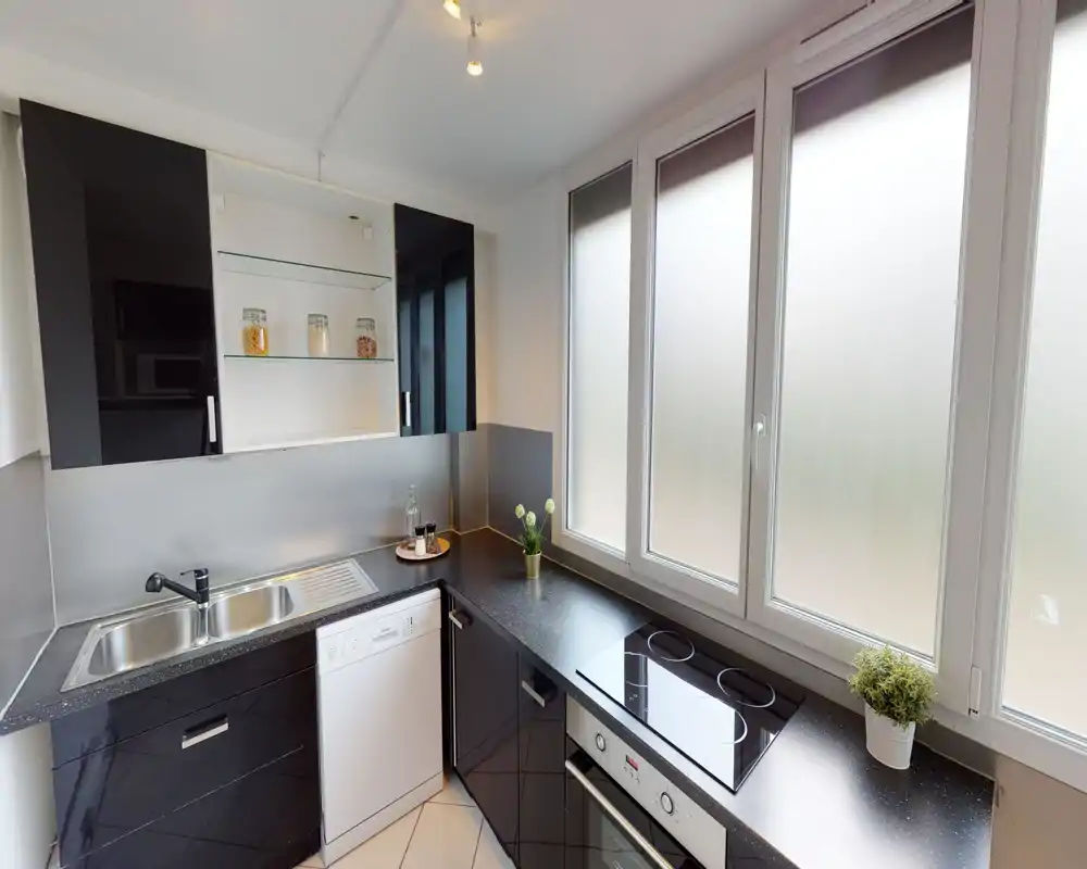 Cucina moderna e luminosa con ante nere lucide, piano cottura a induzione, forno incassato e lavello in acciaio. Ampie finestre smerigliate garantiscono luce diffusa; piano di lavoro ordinato con piccole piante decorative.