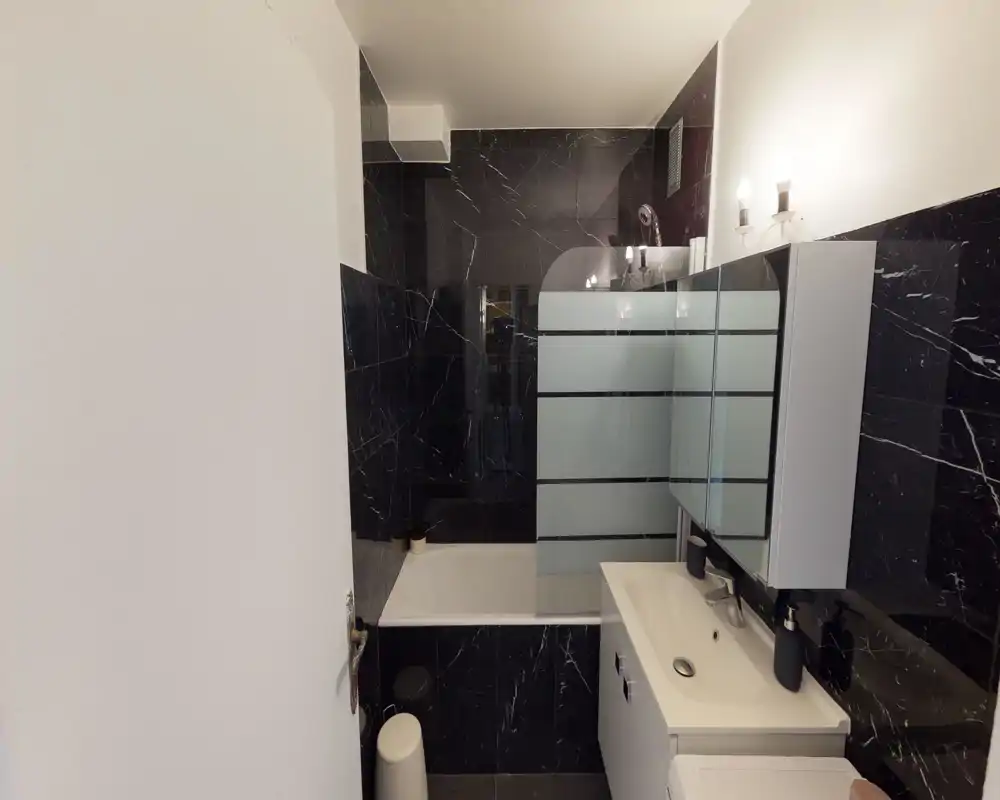 Bagno moderno con vasca, parete doccia in vetro, mobile lavabo a muro e specchiera; piastrelle effetto marmo nero e applique luminose creano un design elegante e contemporaneo.