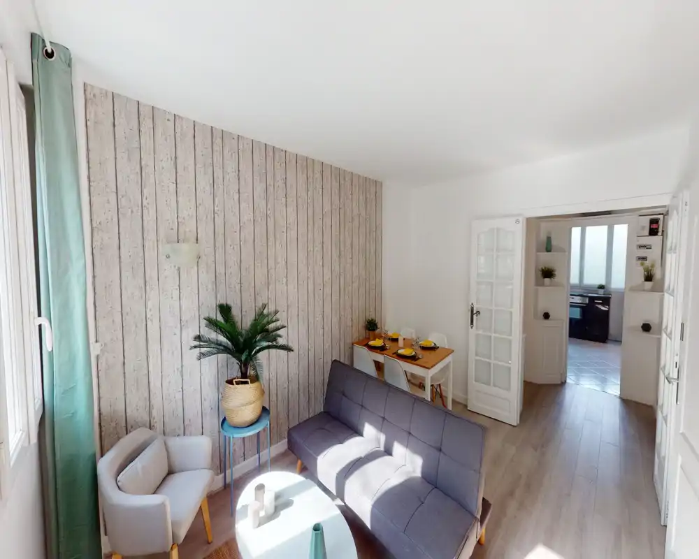 Soggiorno luminoso e ben arredato con divano grigio moderno, una poltroncina, tavolino con pianta e un angolo pranzo compatto vicino alla porta.