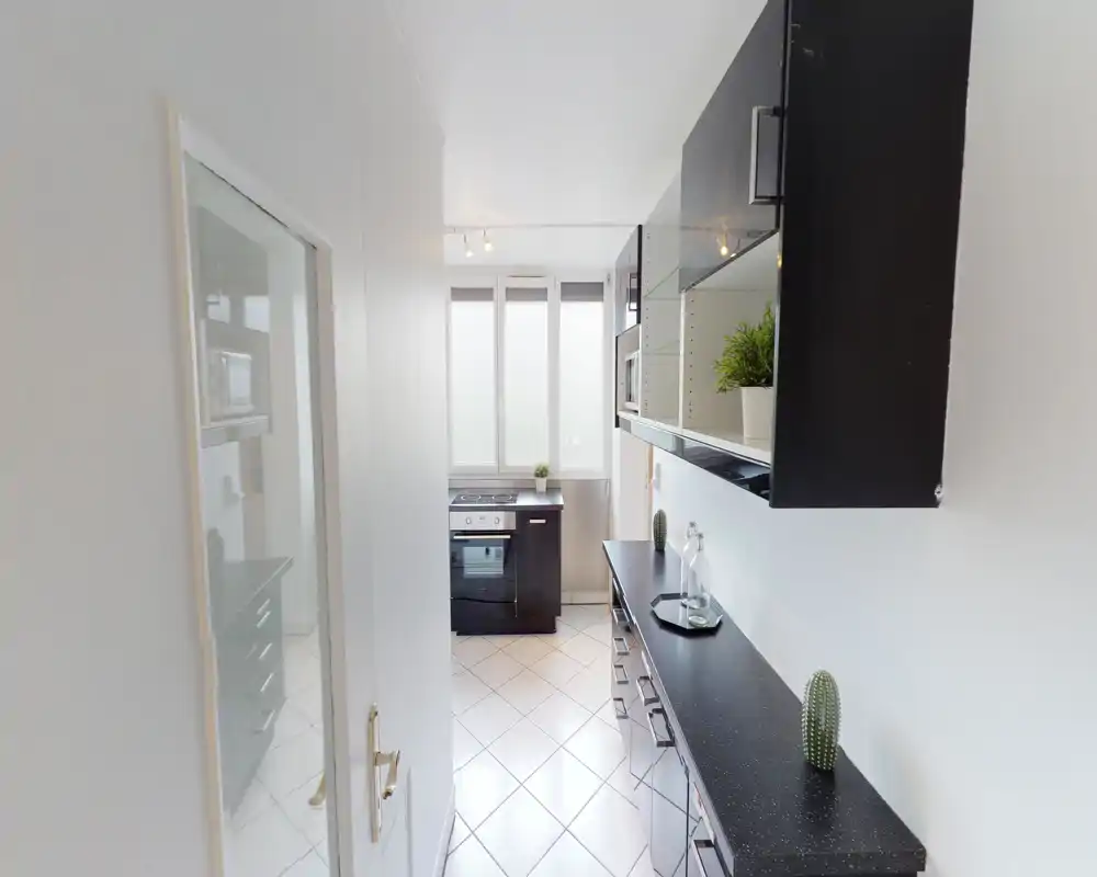 Cucina moderna, stretta e luminosa con pensili neri, forno incassato e ampia finestra che porta luce naturale. Piccoli elementi decorativi come piante e vetri donano un aspetto pulito e accogliente.