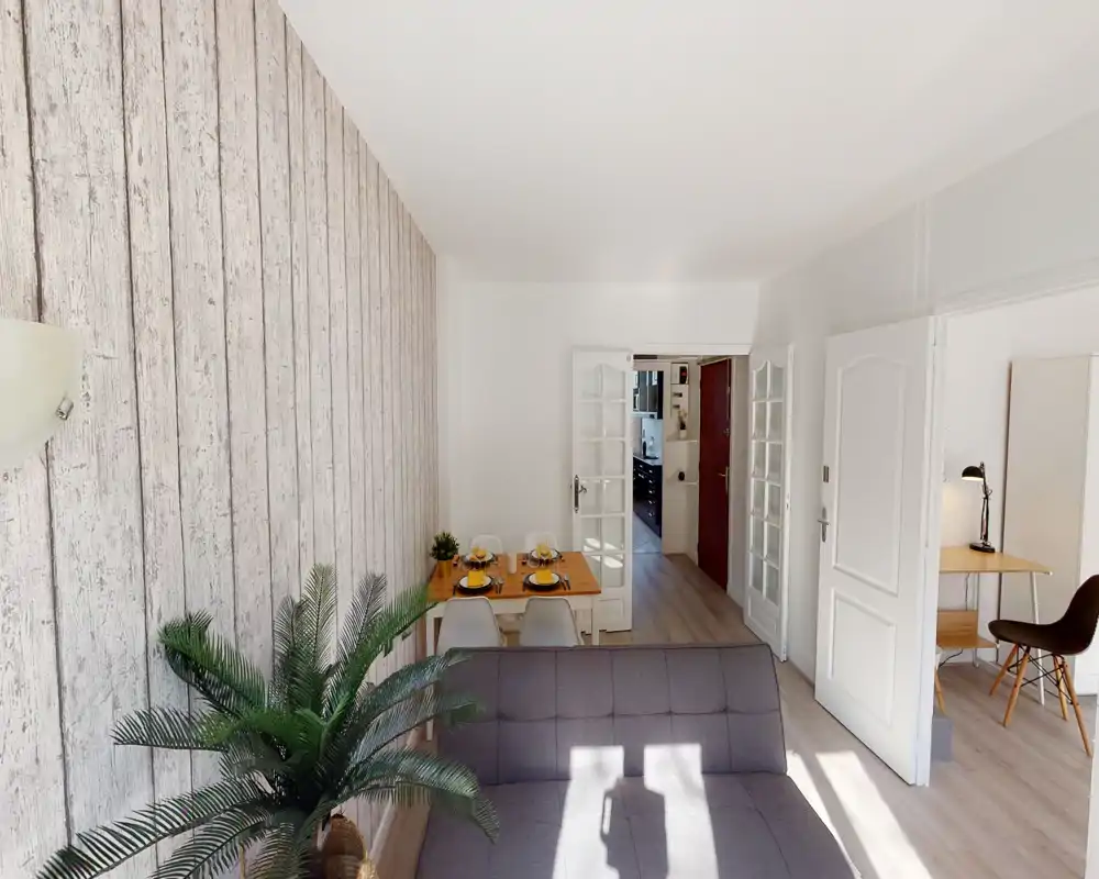 Zona giorno luminosa e accogliente con divano compatto, pianta decorativa e piccolo tavolo da pranzo apparecchiato per quattro; parete effetto legno e luce naturale creano un'atmosfera invitante.