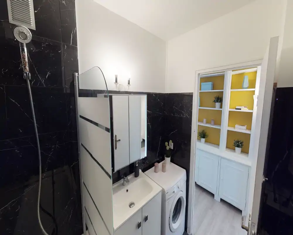 Bagno moderno e luminoso con piastrelle effetto marmo nero, lavabo con mobile specchiato, box doccia in vetro e lavatrice; dalla porta si intravede una libreria nel locale adiacente.