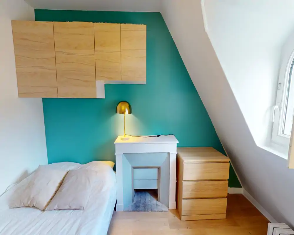 Chambre lumineuse et compacte avec un lit blanc, des oreillers douillets, une lampe de chevet dorée moderne et des rangements en bois devant un mur d'accent bleu-vert.