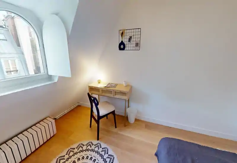 Coin de chambre mansardée avec un petit bureau, chaise, tapis à motif et lit partiellement visible ; une grande fenêtre en arc apporte une belle lumière naturelle.