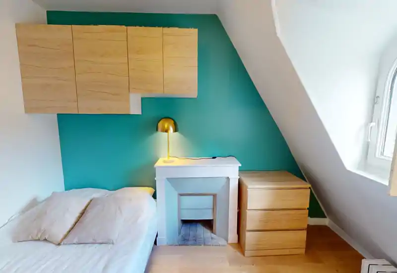 Chambre lumineuse et compacte avec un lit blanc, des oreillers douillets, une lampe de chevet dorée moderne et des rangements en bois devant un mur d'accent bleu-vert.