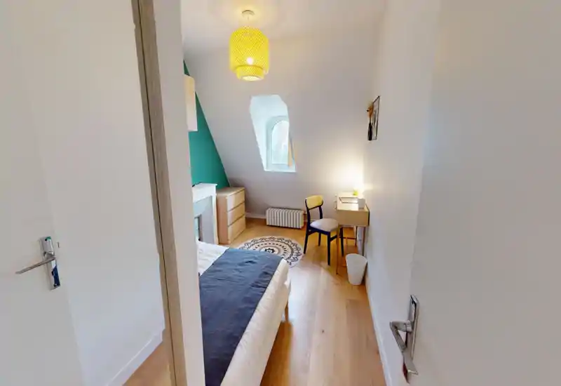 Chambre lumineuse et compacte avec un lit double, parquet, petit bureau et décoration chaleureuse — idéale pour une personne seule ou un couple. Lumière naturelle par une fenêtre en lucarne et lampe d’appoint créent une ambiance agréable.